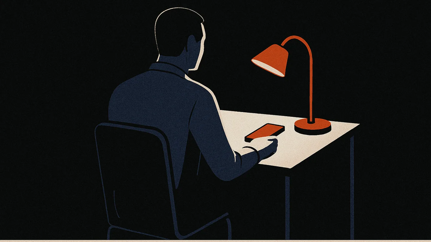 A person at a desk at night holding a phone, lit by a single warm desk lamp.