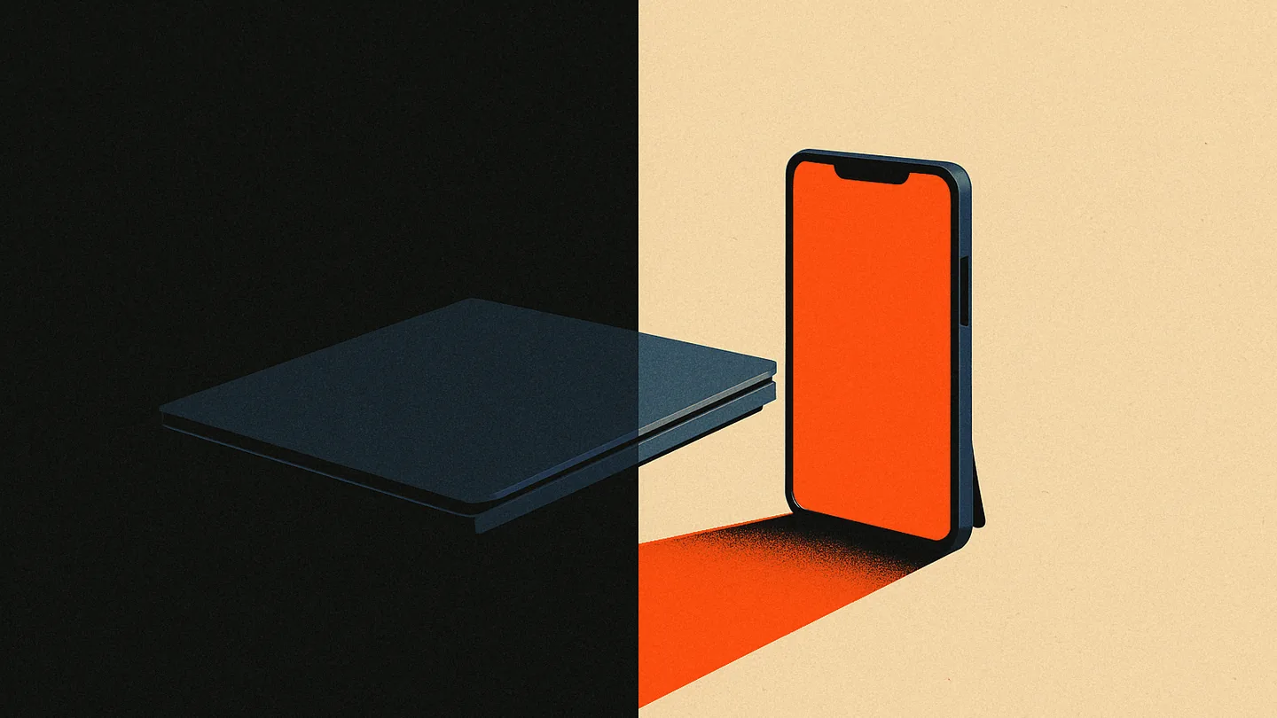 A desk split between a dark, closed corporate laptop and a personal phone with its screen lit orange.