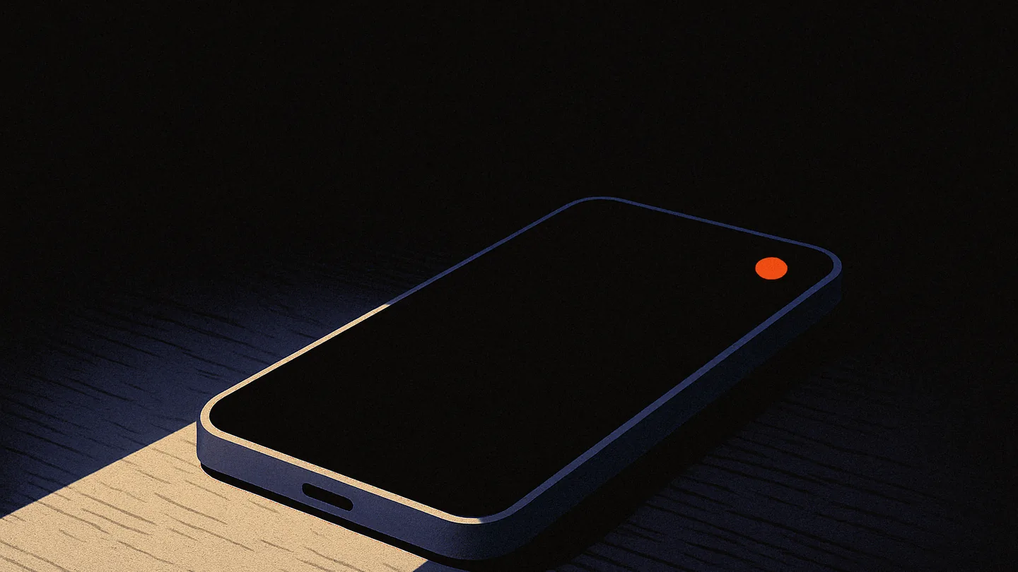 A phone face-up on a dark desk at night, screen dark except for one small orange notification dot glowing in the corner.