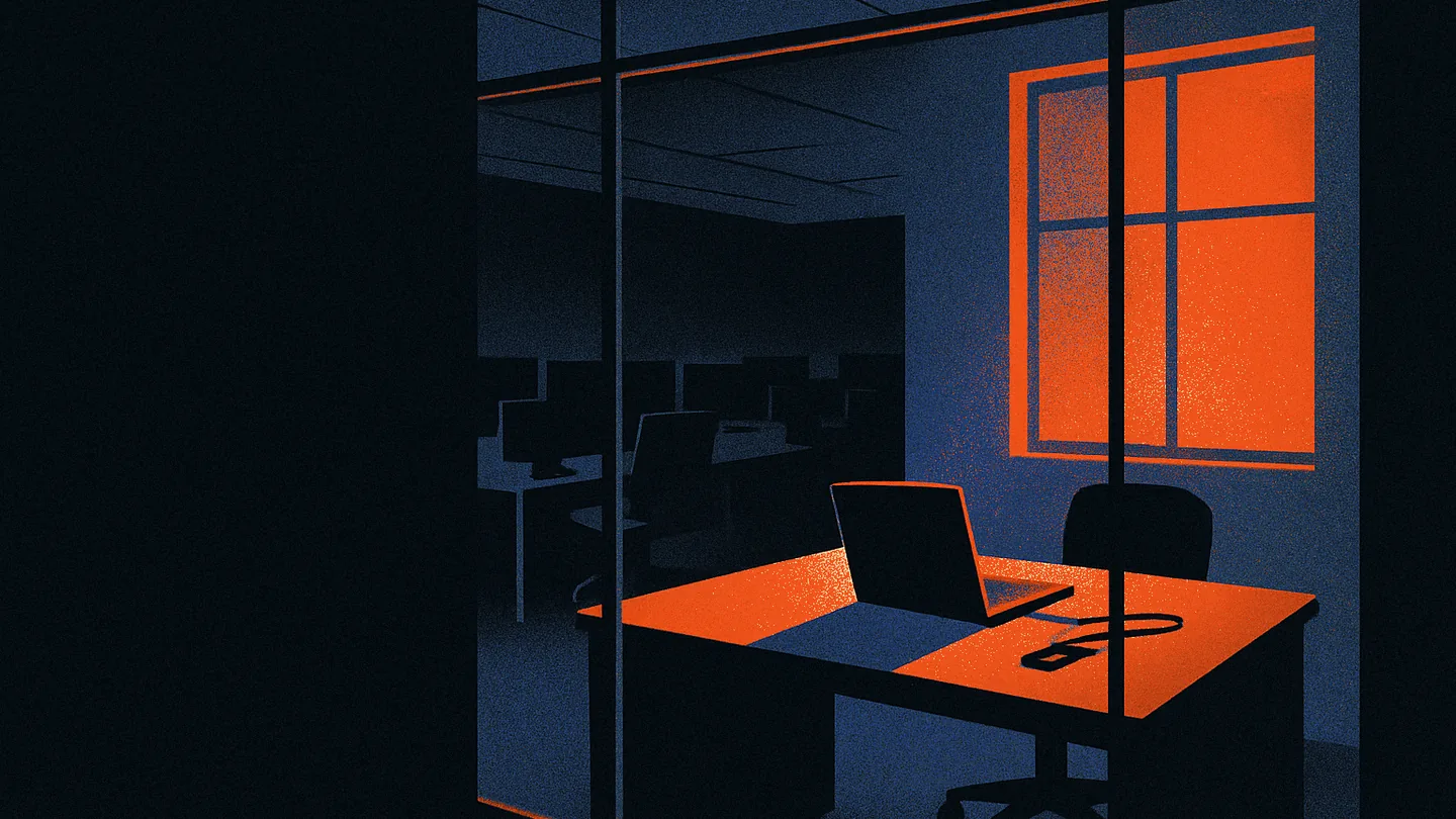 An empty, lit workstation seen through a glass office partition at dusk, a closed laptop with a single glowing orange indicator light.