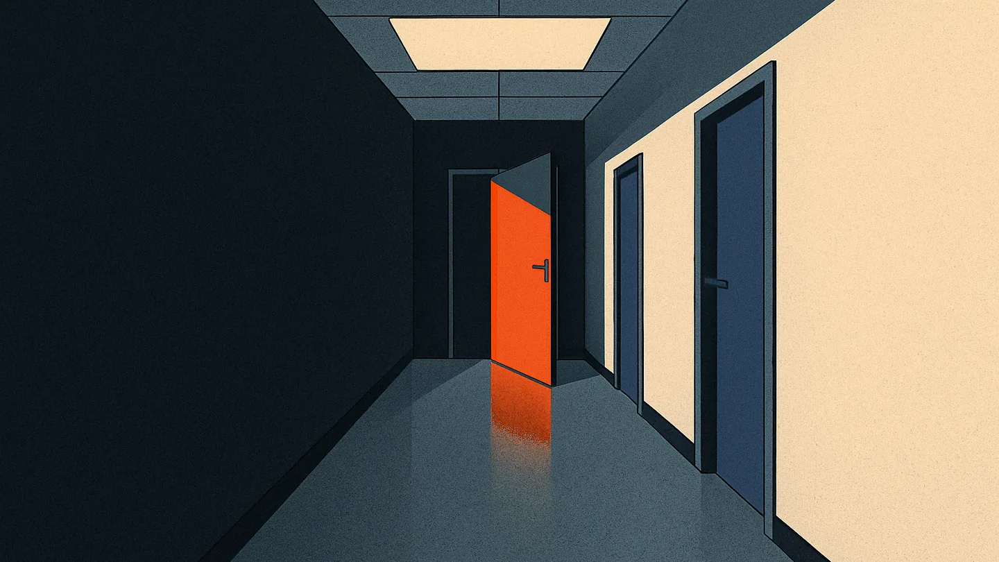 An empty office corridor leading toward a half-open door glowing warm orange at the far end.